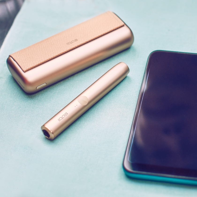 Gold Khaki zlatni IQOS ILUMA PRIME držač i punjač pokraj mobitela.	 	 	 	