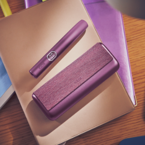 Garnet Red IQOS ILUMA i PRIME device next to its charger, on a pile of notebooks