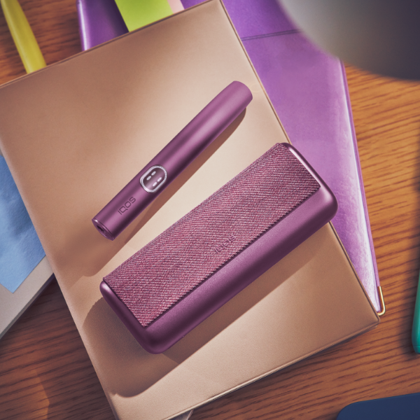 Garnet Red IQOS ILUMA i PRIME device next to its charger, on a pile of notebooks
