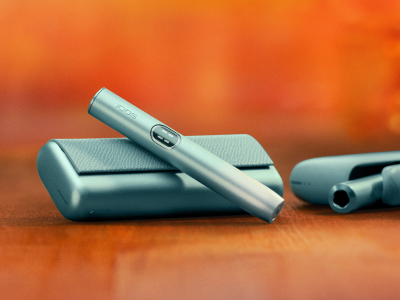 Breeze Blue IQOS ILUMA i PRIME device above its charger, with an IQOS ILUMA device in the background