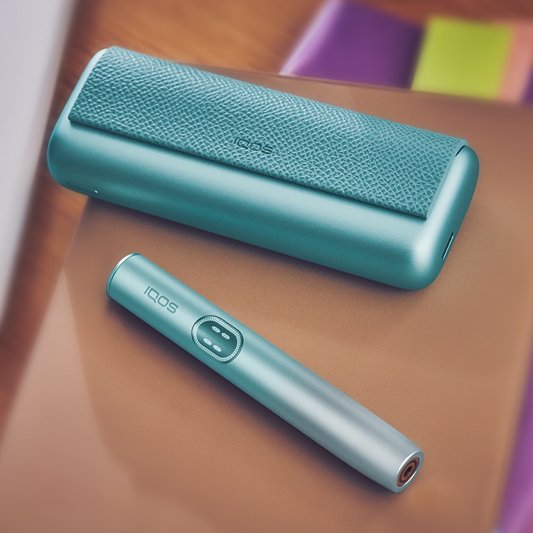 Breeze Blue IQOS ILUMA i PRIME device next to its charger, on a pile of notebooks