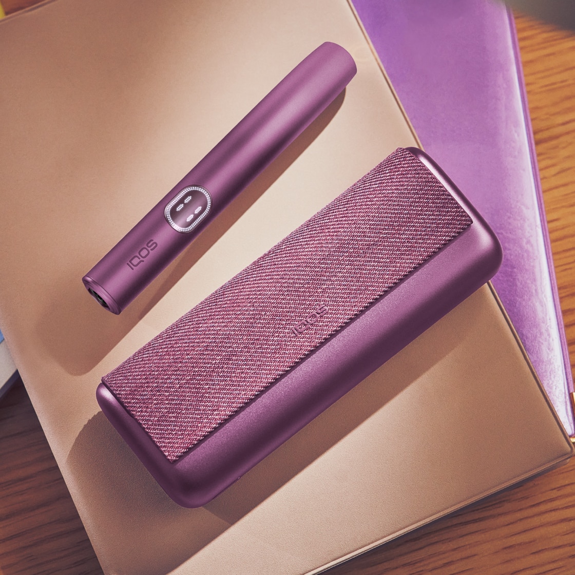 Držač IQOS ILUMA i PRIME uređaja u bordo Garnet Red boji pokraj džepnog punjača, na hrpi notesa